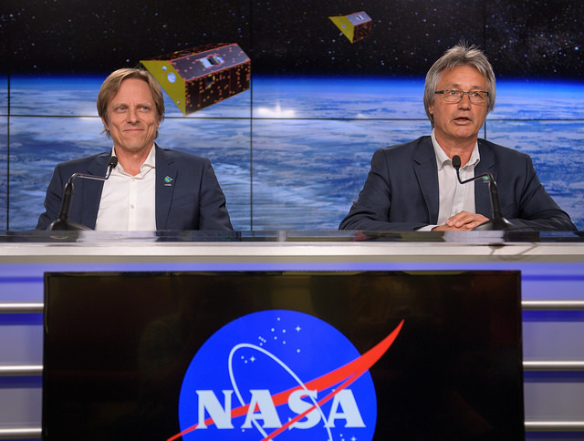 Two scientists sit on a podium with a large NASA logo.