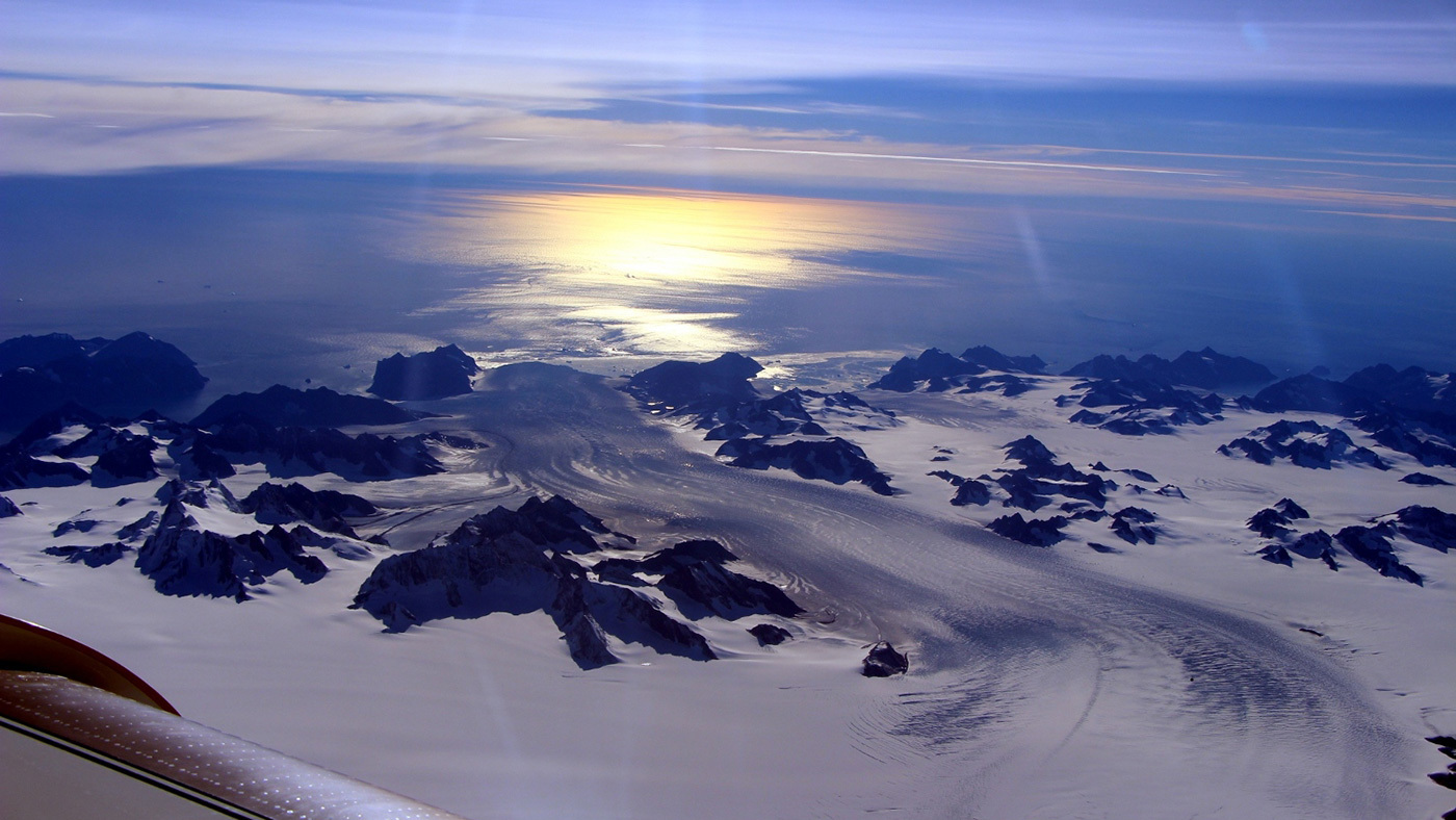 A photo of Steenstrup Glacier from an airplane, with the sun glinting behind it. 